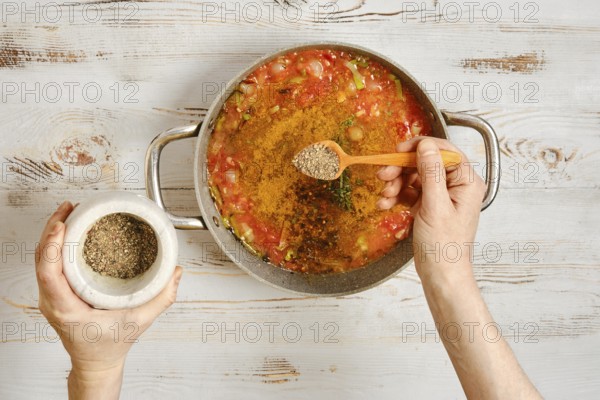 A person adds pepper to a pot of vegetables and tomatoes. The hands hold a wooden spoon and a