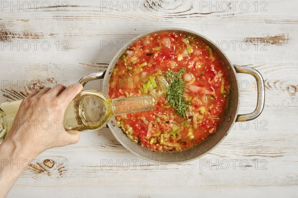 A person pours white wine into a pot filled with chopped tomatoes and vegetables. The pot sits on a