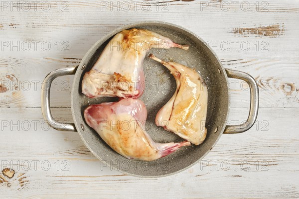 Three raw rabbit legs are browning in a metal pot on a wooden surface. The scene shows the