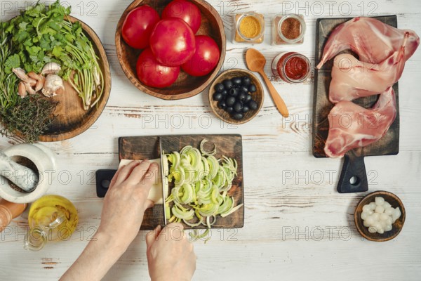 Fresh vegetables, meats, and spices are arranged on a table. Someone is chopping onions with a