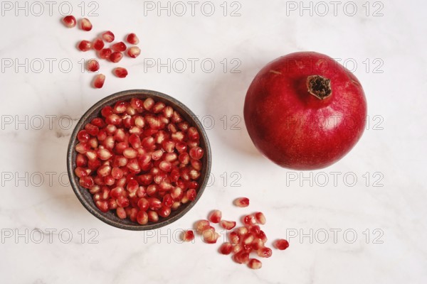 A round red pomegranate sits next to a bowl filled with bright red seeds on a white marble surface.