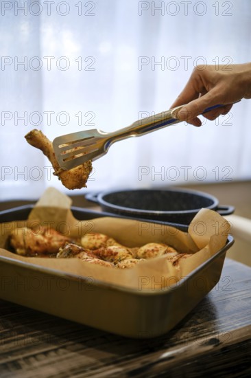 A hand lifts a piece of grilled chicken drumstick from a tray. The scene shows a cozy kitchen setup