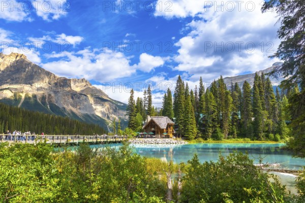 Wooden lodge reflecting on turquoise water with tourists walking on a bridge, surrounded by