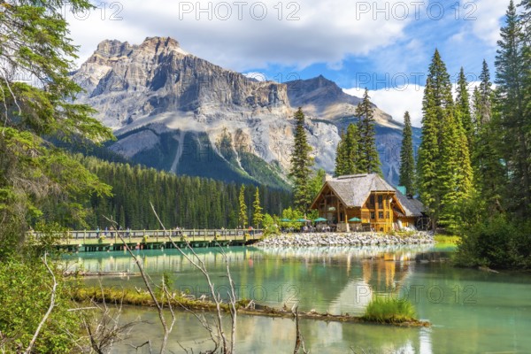 Wooden lodge reflecting in turquoise water on sunny summer day with tourists walking on bridge and