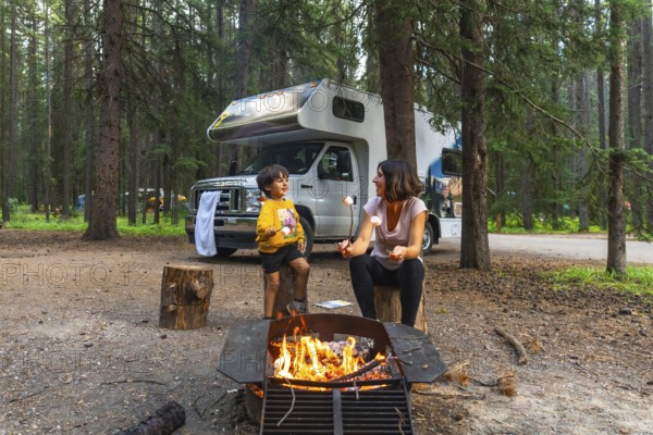 Mother and son roasting marshmallows over campfire near their camper van, enjoying a camping trip