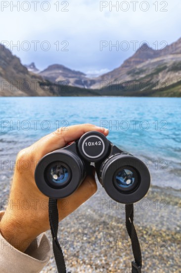 Hand holding binoculars, focusing on turquoise waters of bow lake surrounded by majestic mountains