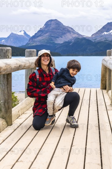 Mother happily holds her son on a wooden bridge, enjoying the stunning turquoise waters of bow lake