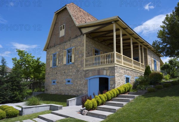 Traditional stone house with veranda and shutters, surrounded by well-kept garden under blue sky,