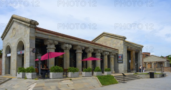 Historic stone building with ancient columns and red umbrellas under cloudy sky, Mtskheta,