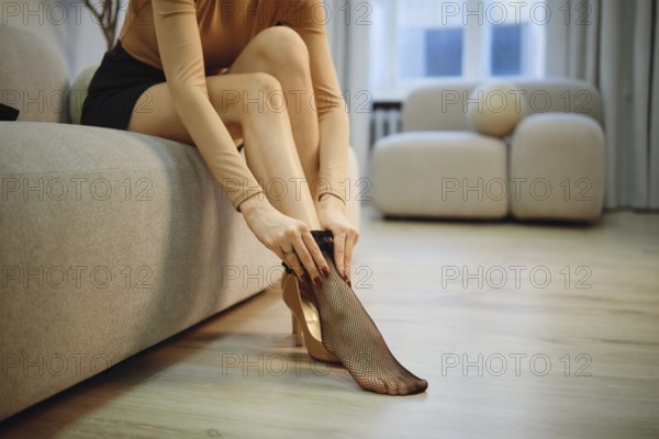 Unrecognizable woman is seated on a couch in a bright room. She is putting on black fishnet