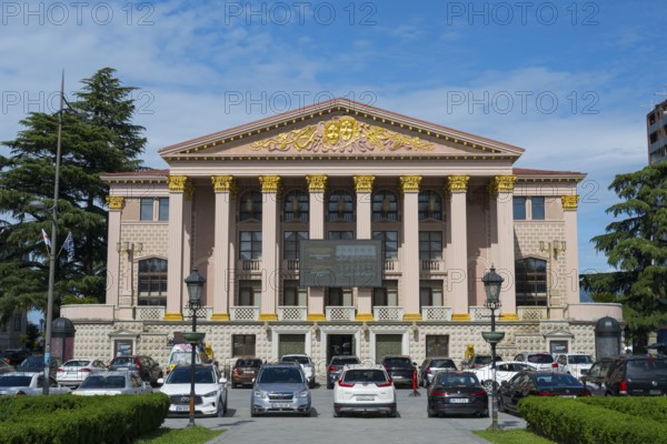 An imposing building with ancient columns and golden decoration, cars parked in front of it, Ilia