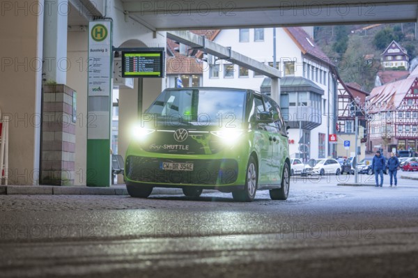 Green shuttle at a covered stop, surrounded by half-timbered houses, Deer E-Mobility Shuttle