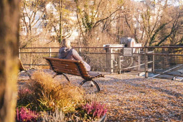 Woman sitting on park bench looking at river and prohibition sign in autumn, Stadtgarten Pforzheim,