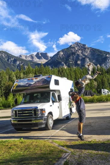 Father playfully lifting his son in the air in front of their camper van, enjoying a scenic stop
