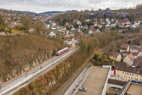 A train travels through a hilly area with urban houses and winter trees under a cloudy sky,