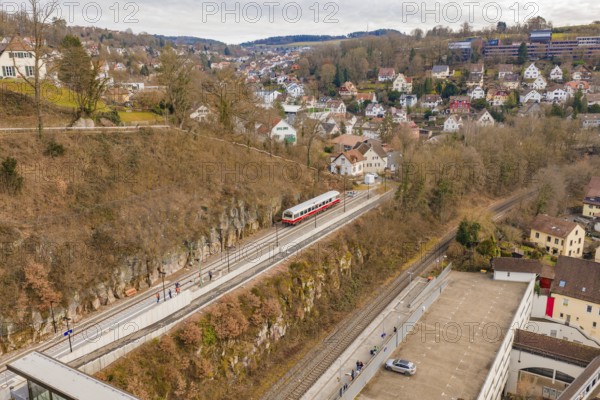 A train moves through an urban environment with hills and houses, surrounded by winter vegetation