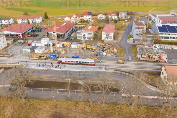 A construction site next to a train, surrounded by buildings and fields, is viewed from the air,