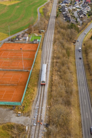 A train travels along railroad tracks past tennis courts, next to a busy road, historic event,