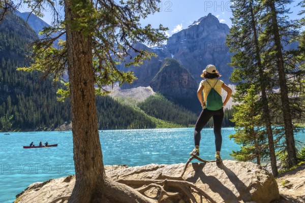 Female tourist standing on a rock by a tree, admiring the turquoise waters of moraine lake with
