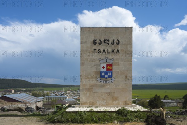 A monument with the coat of arms of Tsalka against a village backdrop and a blue sky, town sign