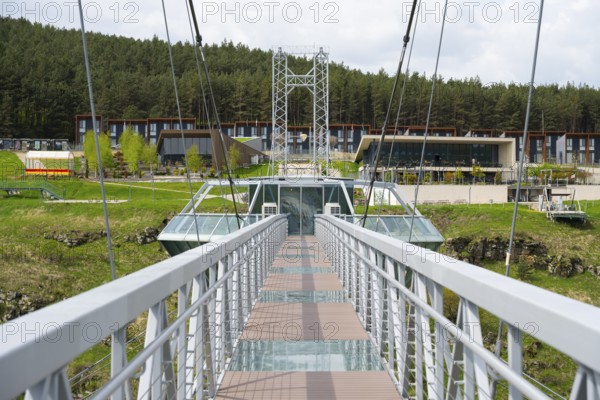 View of a modern suspension bridge with buildings in the background and surrounded by forest,