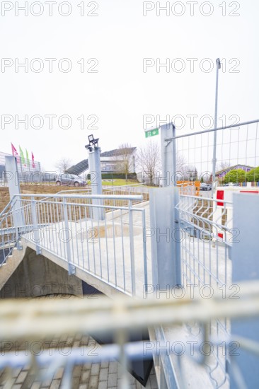 Modern outdoor area with steel railings and flags, building complex in the background, parking