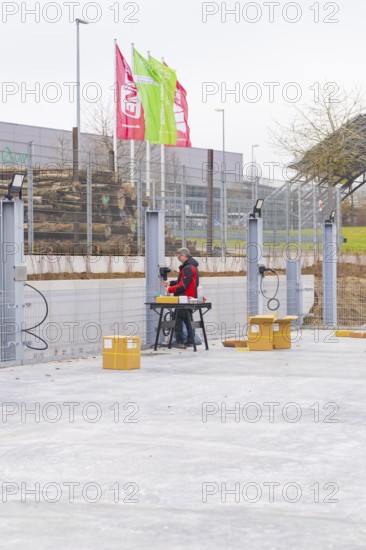 A worker sitting at a table next to a fence with assembled cables and boxes in an industrial area,