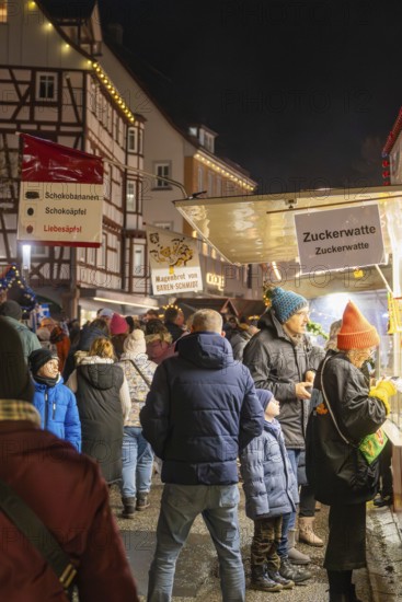 Night market visitors at an illuminated cotton candy stand in front of half-timbered houses,
