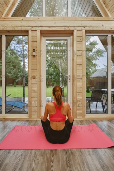 Back view of a woman sitting cross-legged on a pink yoga mat inside a wooden cabin. Outside, there