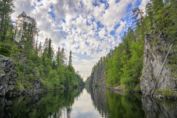 A calm lake lies in a wooded gorge, surrounded by tall rocks and reflecting the sky covered with