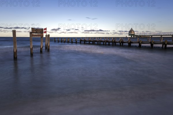 Wooden sign in the sea with inscription Zingst and wooden walkway, pier with diving gondola behind,