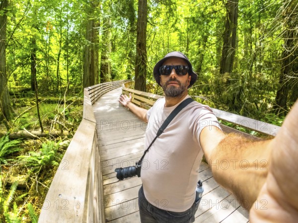 Photographer with hat and sunglasses taking a selfie while walking down a wooden walkway in a lush