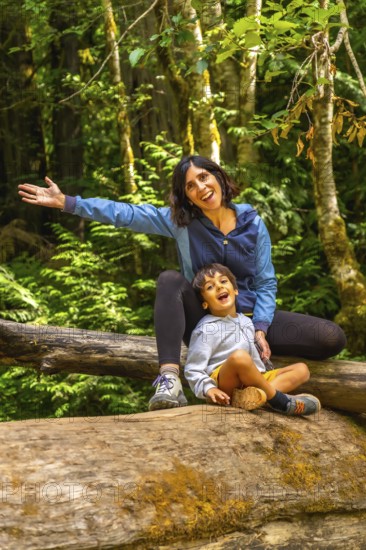 Cheerful mother and her son are sitting on a large fallen log in cathedral grove forest on