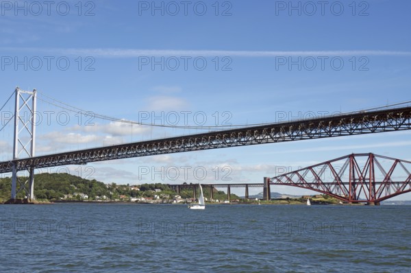 Two distinctive bridges over water with a small sailboat in the foreground, railway bridge and road