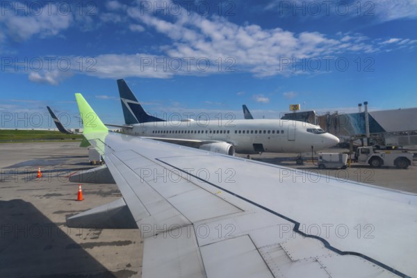 Airplane wing is overlooking airport runway with other aircraft parked and ground crew servicing