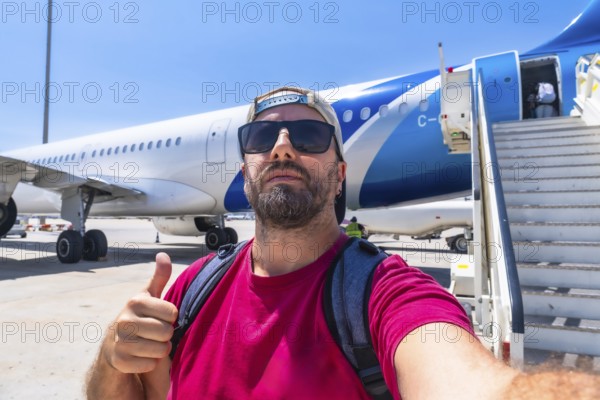 Happy bearded tourist wearing sunglasses and backpack taking selfie showing thumbs up gesture while