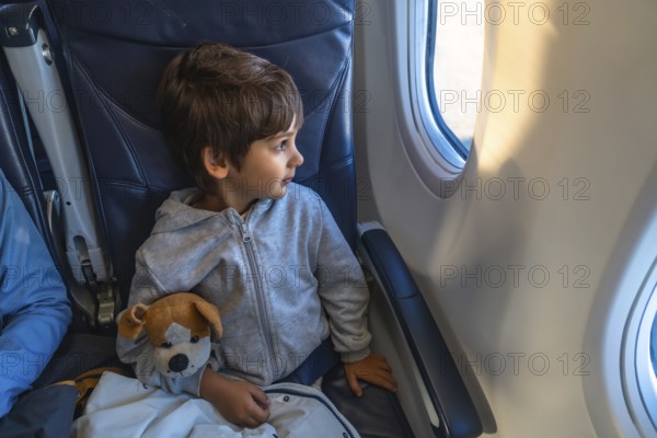 Curious child traveling by plane, clutching a plush toy while gazing out the window, marveling at