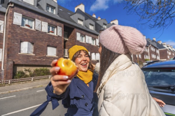 Two young women laughing and sharing a healthy snack on a sunny urban street, bundled in coats and