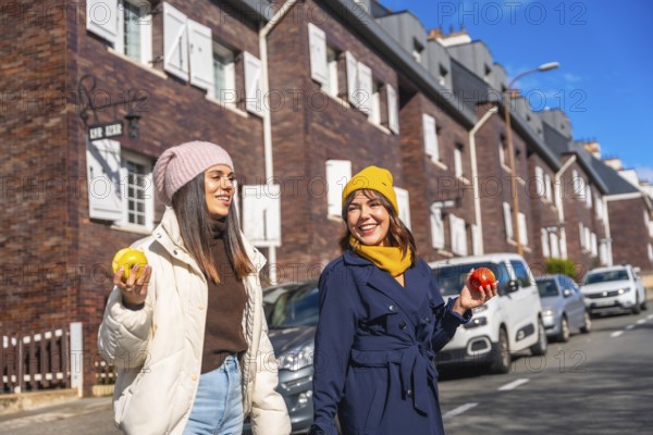 Two cheerful young women enjoying a healthy snack of apples while walking and smiling on a city