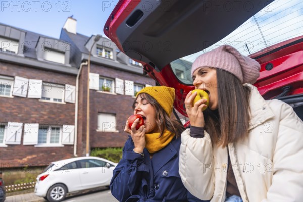 Two young women sit in an open car trunk on a city street, laughing and sharing fresh apples during