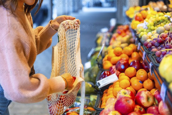 Woman selecting organic apples and oranges, placing them into a sustainable net shopping bag while