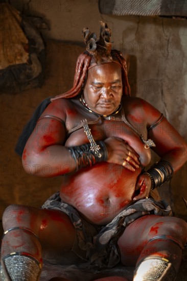 Elderly Himba woman rubbing herself with red paste for skin care, in a hut, traditional Himba