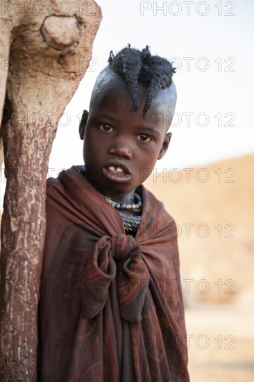Himba girl leaning at a traditional clay hut, in the morning, traditional Himba village, Kaokoveld,