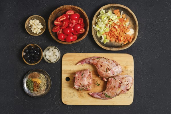 A rabbit's hind legs and a saddle are placed on a wooden cutting board, surrounded by fresh