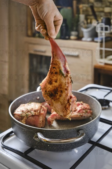 A person holds a marinated rabbit hind leg above a pot with other meat cooking inside. The kitchen