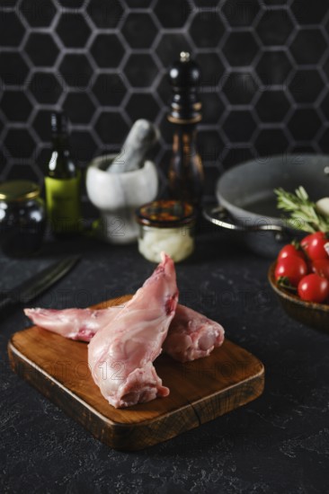 Raw rabbit hind legs on a wooden cutting board in a well-equipped kitchen. Fresh vegetables, oils,