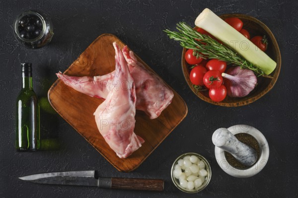Top view of two raw rabbit hind legs are on a wooden board surrounded by fresh vegetables, herbs,