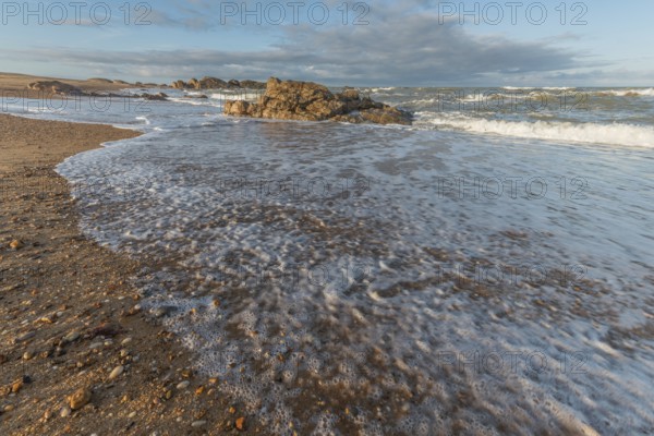 A wide sandy beach stretches out with scattered pebbles and beach shells. Gentle waves crash