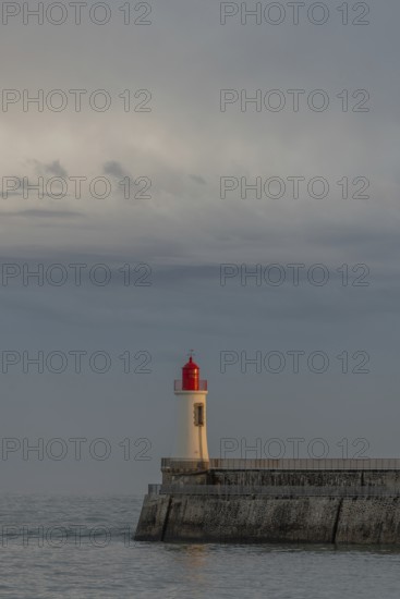 A lighthouse stands on a pier and casts its light over calm waters at dusk. The sky is filled with
