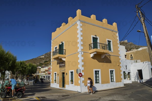 Mediterranean-style corner building with balconies and passers-by under a clear blue sky, Agia